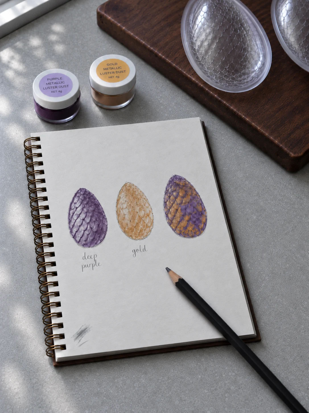 An open sketchbook on a kitchen worktop showing three painted dragon egg designs in purple, gold and the two mixed, beside jars of lustre dust
